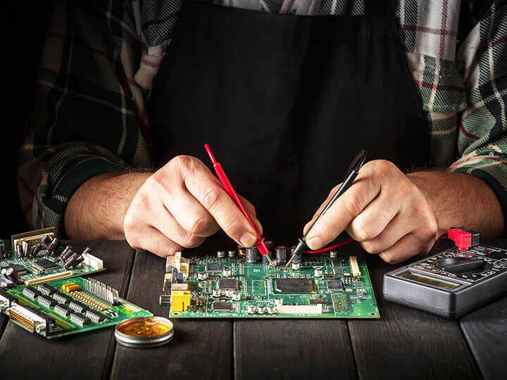 Electrician in a plaid shirt and black apron testing a circuit board assembly with an electric multi-tester tool